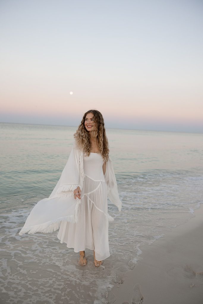 Alyssa, founder of HolSol, walking through shallow waters at sunset, wearing all white