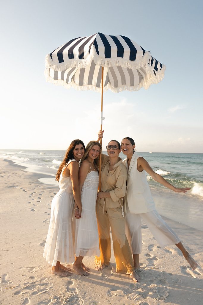 4 women, all part of the HolSol team, stand smiling at the beach under a striped umbrella that is hoisted in the air