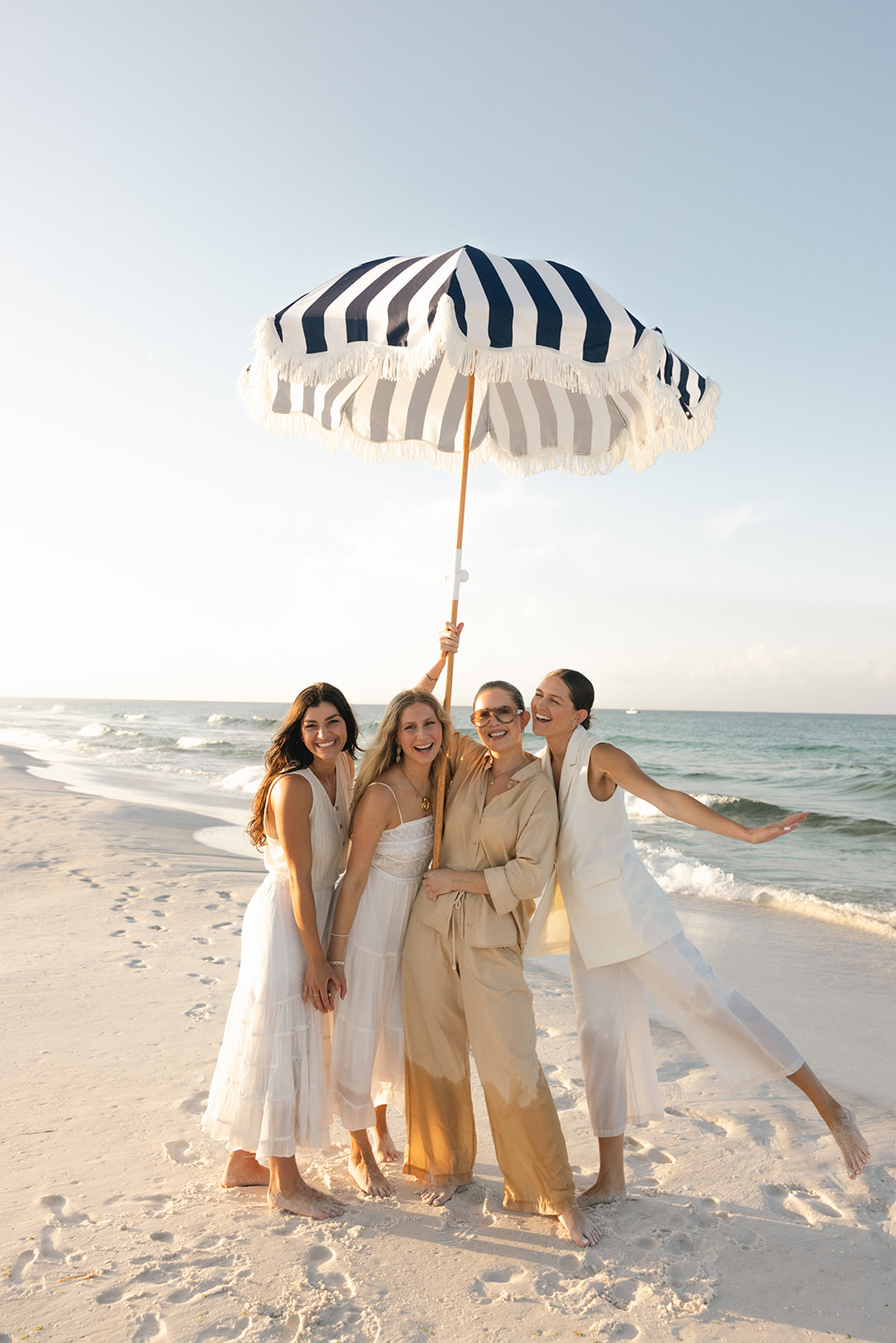 4 women, all part of the HolSol team, stand smiling at the beach under a striped umbrella that is hoisted in the air
