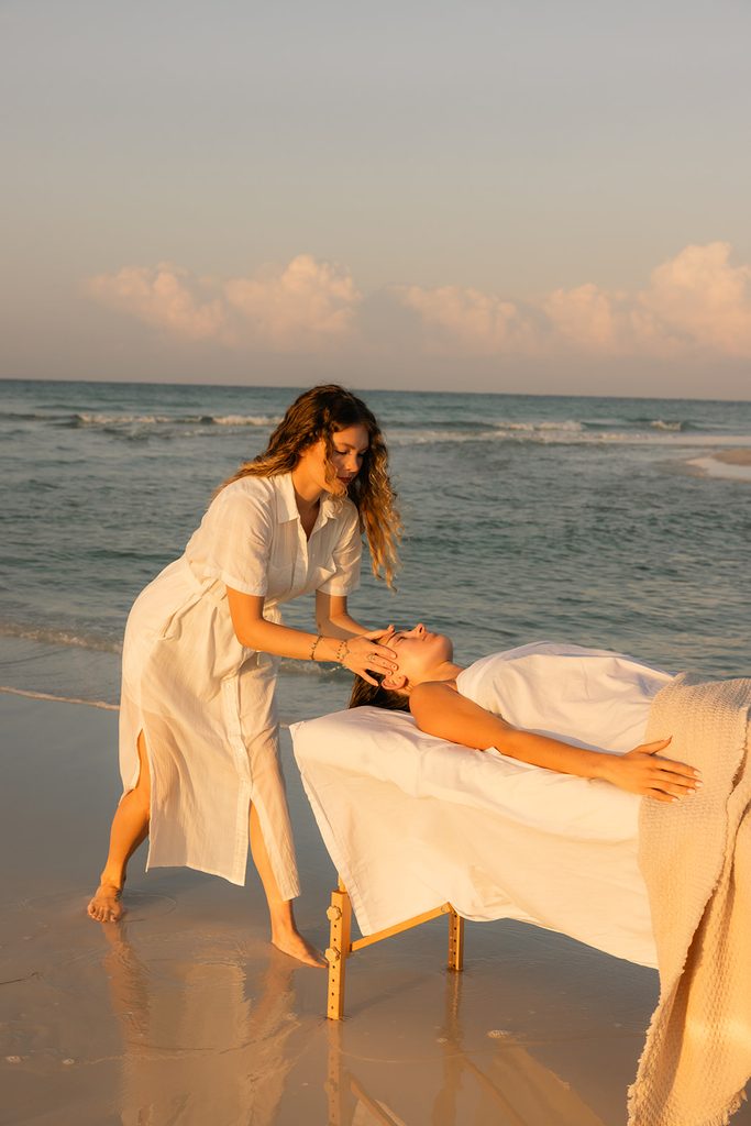 Alyssa, founder of HolSol, giving a client a facial at the beach, near the shallow waters, during golden hour